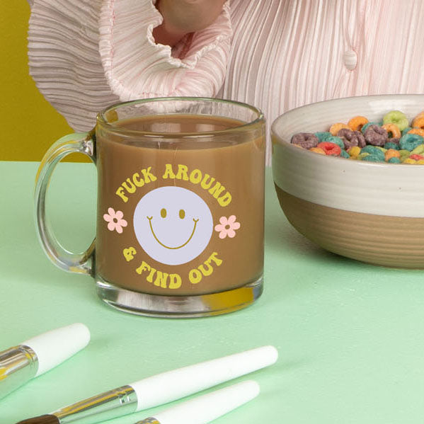 Coffee in the citron "fuck around and find out" text glass mug with a blue smiley face and small daisys on the each side with a bowl of cereal on the side on a mint color surface 
