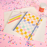 Rainbow checkered notebook with college rule pages and pink, green, and coral pen on top displayed on a pink background with confetti around. "You're doing great" candle in the back.