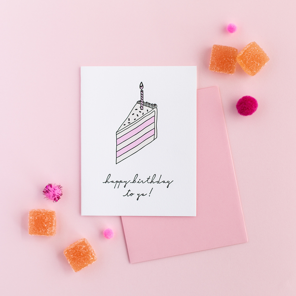A white greeting card with a wedge slice of yellow and pink cake with a single candle on top. There is a pink envelope, orange candy and pink poms against a pink background.