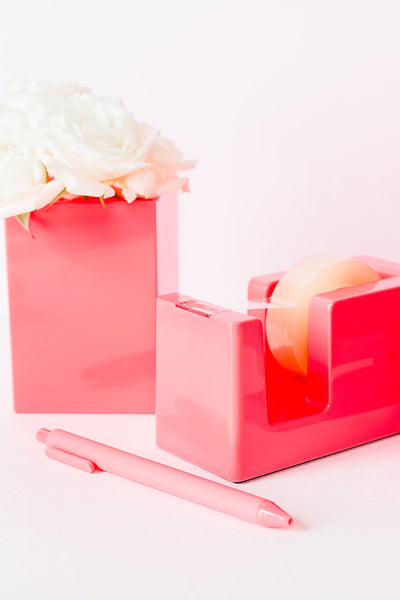 Lifestyle photo of neon coral tape dispenser, pen, and pen cup with flowers in it.