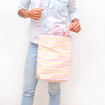 A woman stands in a washed denim shirt with a pink and yellow striped crossbody tote to her side. 