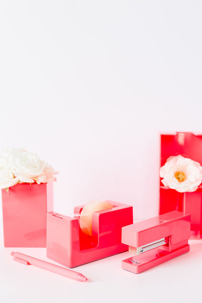 Lifestyle photo of neon coral tape dispenser, tray,  pen, and pen cup with flowers in it.