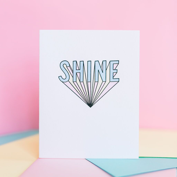 White greeting card standing open with 3d text "SHINE" in blue pink and yellow. There are multiple envelopes in assorted pastel colors under the card. The wall in the background is pink.