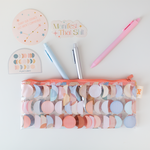 Many Moons Multi Pixie Pouch with moon phase designs in pastel blue peach blush sitting on a white desk with astrology and zodiac stickers, pastel and metallic colored jotter pens