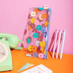 Clear daisy pixie pouch along with jotter pens and other misc items on an orange surface and pink background.