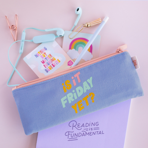 Canvas Pencil Pouch in purple with colorful saying "is it Friday yet?" and a peach zipper. Open with headphones and desk accessories spilling out on top of light purple notebook with "reading is fundamental" on cover.