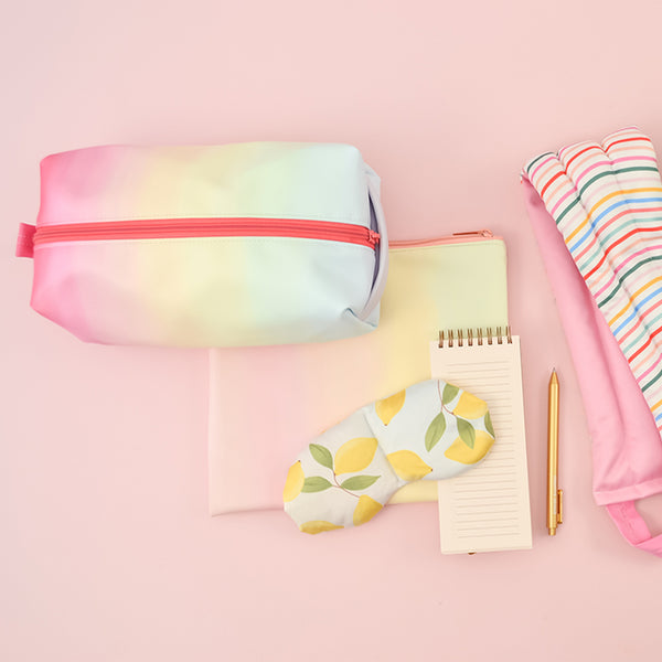 Meltdown Doppelganger is a rainbow print with a coral zipper and carrying handle. Displayed with and eyemask, a Daybreak Pouch, a To-do task pad, a Gold Jotter Pen, and a weighted Neck Wrap.