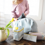 Blonde woman in a pink sweater rummaging through a blue overnight bag, with several makeup pouches and toiletry bags on the table.