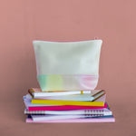 Daybreak Tweedle Dee is a cute cosmetics bag in peach with a rainbow pastel detail shown sitting on a stack of notebooks.