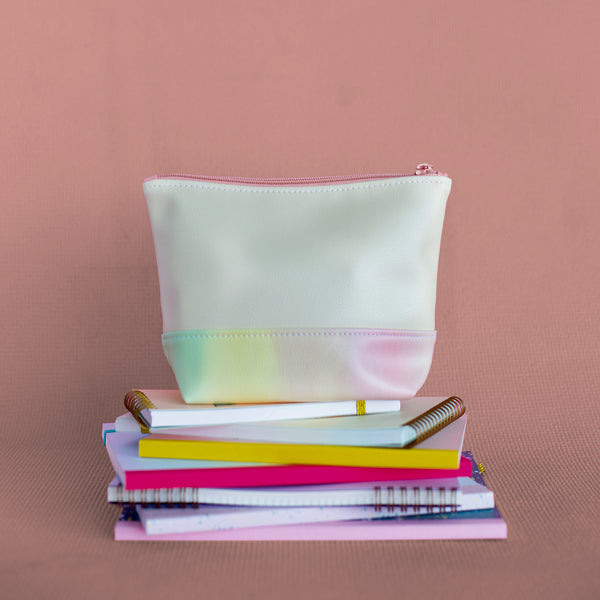 Daybreak Tweedle Dee is a cute cosmetics bag in peach with a rainbow pastel detail shown sitting on a stack of notebooks.