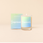 Candle rocks glass with sea foam green-to-light blue ombre decal and text that reads "Capricorn" with minimalist, white sparkle stars surrounding the text; "the detailed over-achiever" sits at the bottom of the decal. Box packaging with the same design sits behind glass.