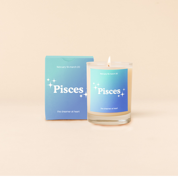 Candle rocks glass with light-to-dark blue ombre decal and text that reads "Pisces" with minimalist, white sparkle stars surrounding the text; "the dreamer at heart" sits at the bottom of the decal. Box packaging with the same design sits behind glass.