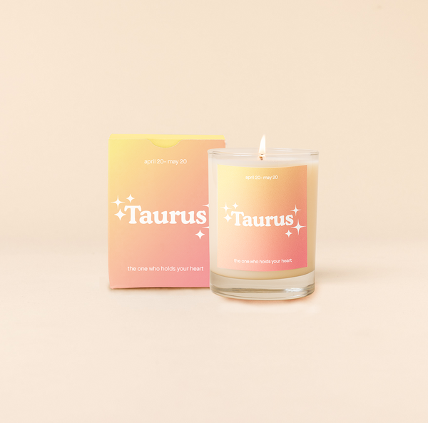 Candle rocks glass with yellow-to-orange ombre decal and text that reads "Taurus" with minimalist, white sparkle stars surrounding the text; "the one who holds your heart" sits at the bottom of the decal. Box packaging with the same design sits behind glass.