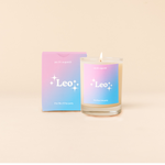Candle rocks glass with pink-to-bright blue ombre decal and text that reads "Leo" with minimalist, white sparkle stars surrounding the text; "the life of the party" sits at the bottom of the decal. Box packaging with the same design sits behind glass.