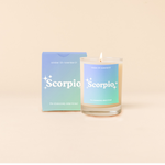 Candle rocks glass with light blue-to-light green ombre decal and text that reads "Scorpio" with minimalist, white sparkle stars surrounding the text; "the obsessively determined" sits at the bottom of the decal. Box packaging with the same design sits behind glass.