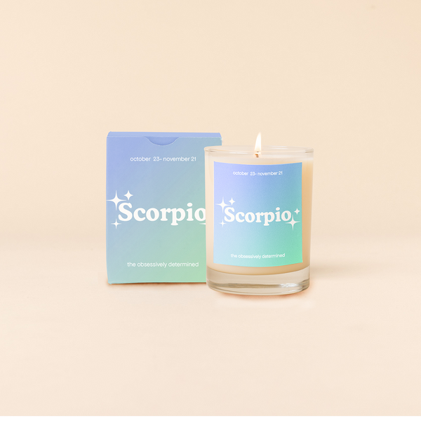 Candle rocks glass with light blue-to-light green ombre decal and text that reads "Scorpio" with minimalist, white sparkle stars surrounding the text; "the obsessively determined" sits at the bottom of the decal. Box packaging with the same design sits behind glass.