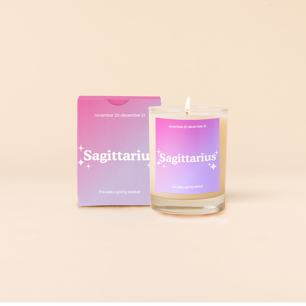 Candle rocks glass with dark pink-to-lavender ombre decal and text that reads "Sagittarius" with minimalist, white sparkle stars surrounding the text; "the easy-going seeker" sits at the bottom of the decal. Box packaging with the same design sits behind glass.
