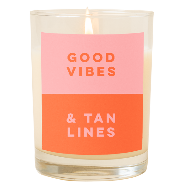 A candle with a decal that says, "Good vibes and tan lines." Top half is a dusty pink color and bottom half of decal is orange.