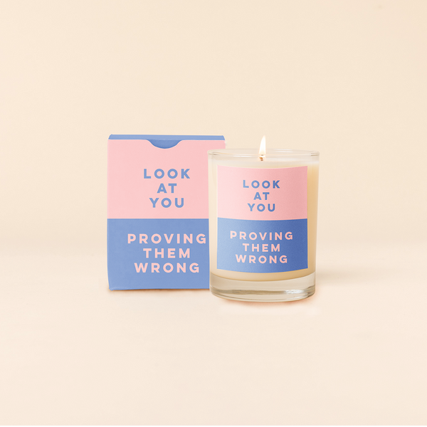 Rocks candle with two tone backdrop. Top half pink backing with blue text and bottom half blue backing with pink text reading "LOOK AT YOU PROVING THEM WRONG". Two tone box packing with same text as candle.