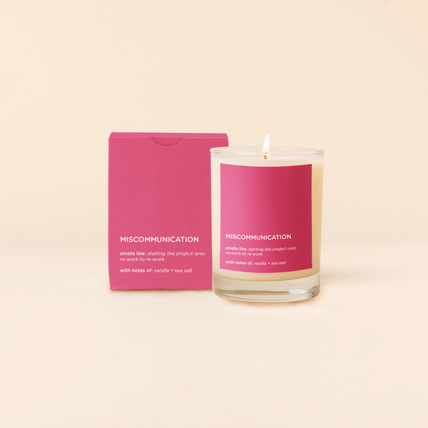14 oz candle rocks glass with pink decal and text that reads "MISCOMMUNICATION, smells like:starting the project over. re-work to re-work. with notes of: vanilla + sea salt" in white font. Pink box packaging with same design sits behind candle.