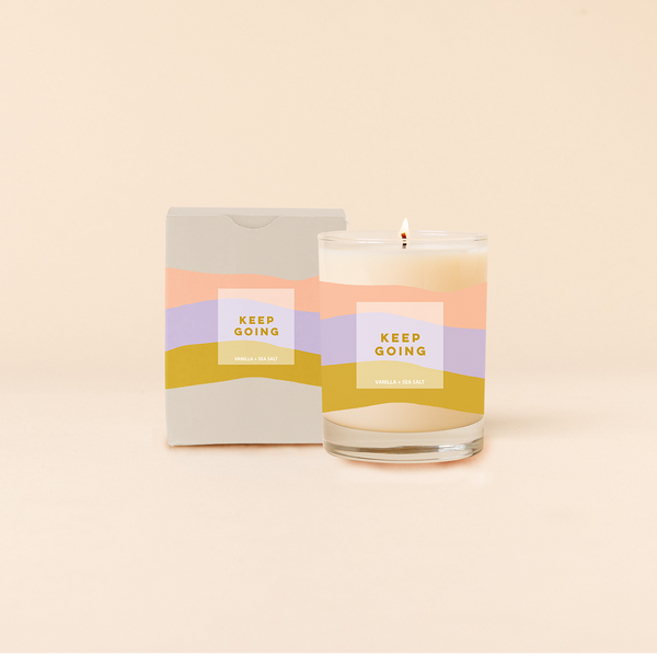 Rocks candle reading "KEEP GOING" in front of tri-color horizontal stripes around candle. Box packaging with same design and text as candle. 