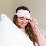 A woman wearing a Not Today sleep eye mask. Mask is a blush pink with multicolored letters that say "Not Today" and the perimeter of the eye mask is multicolored mini hearts.