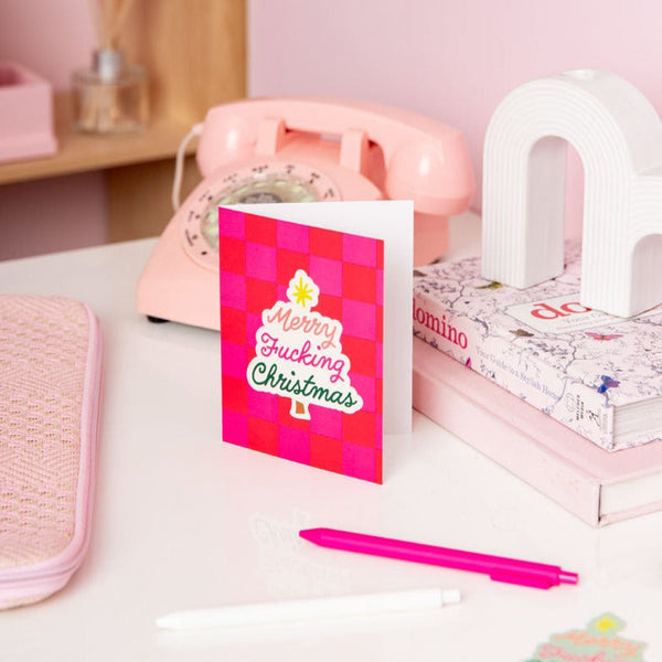 Hot pink and red checkered "Merry Fucking Christmas" card on a white desk with pink items.