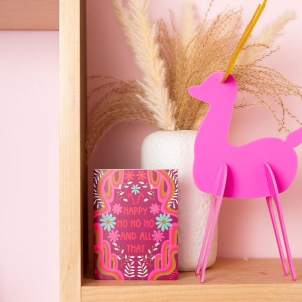 A hot pink reindeer decoration standing next to a red and pink "Happy ho ho ho and all that" greeting card.