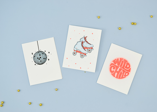 A collection of white greeting cards. From left to right: A light blue and black disco ball. A light blue and coral pair of skates. A coral and red circular graphic with "Wild Child".