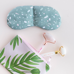 A weighted eye mask in a green terrazo print is sitting next to a light pink vegan leather zippered pouch. The pouch has a green leaf print all over it and has two stone face rollers tucked into it. 