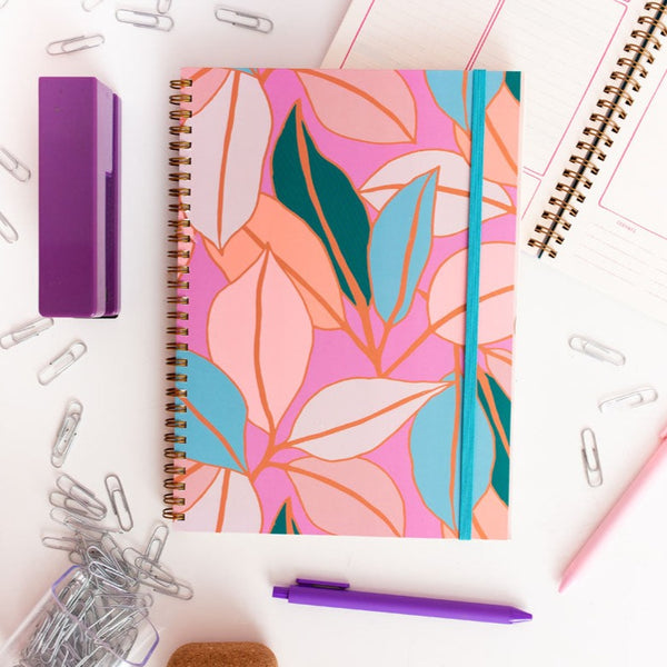 small pink goal getter with multicolor pastel leaves and a blue elastic band to close it. surrounding the planner are paper clips, pen jotters and a purple stapler.