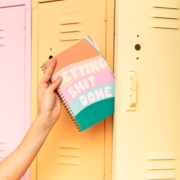 a girl putting in a small goal getter planner with five waves in green, blue, pink, peach and orange, the words 'getting shit done' in white fluffy text at the front.