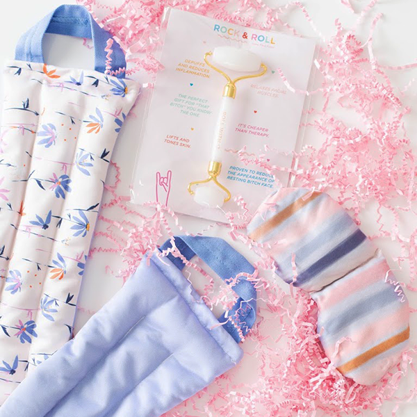 Contents of a gift set laid out to show the floral patterned neck wrap, a weighted eye mask with blue stripes printed on it, and a white quarts face roller amidst pink paper shreds.