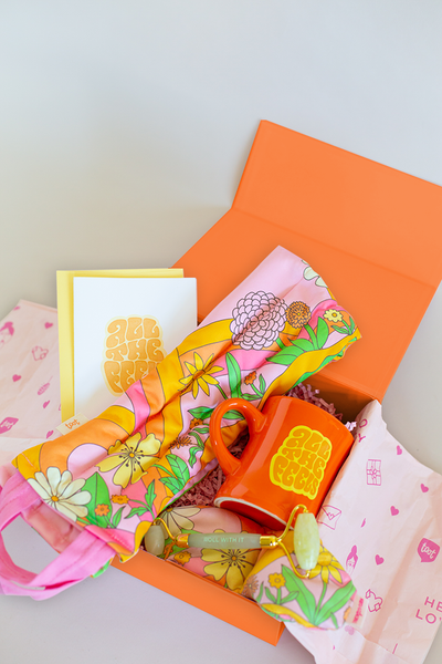 a collection of self care items packaged in an orange box including a neck wrap and an eye mask in a pink floral and swirl print, an orange diner style mug and  a card that say all the feels, and a face stone roller 