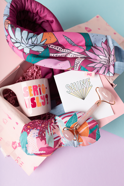 A Floral nights selfie care kit with a pink "Seriously" mug, a "Floral Nights" eye mask, a "Floral Nights" weighted neck wrap, a Rose Quartz face roller, and a greeting card that says "SHINE" in dusty blue letters.  Displayed in gift box.