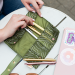Woman placing Jotter Pens inside of An olive green roll up pouch with stencil women faced in white and pink. Pouch is good for holding brushes, pens, pencils, small electronics and has buttons to close the pouch after rolling up.