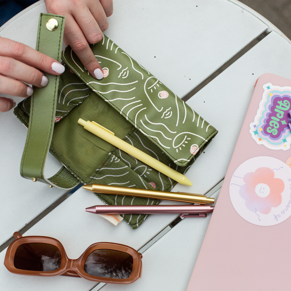 Woman placing Jotter Pens inside of An olive green roll up pouch with stencil women faced in white and pink. Pouch is good for holding brushes, pens, pencils, small electronics and has buttons to close the pouch after rolling up.