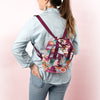 A jewel-toned backpack with a front pocket. Designed with succulents, rainbow arches and abstract lines. Displayed by person holding bag in front of pink background.