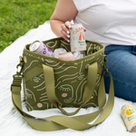 woman in jeans and white shirt sitting on a white picnic blanket with olive green ice princess cooler with white silhouette of a woman's face with pink cheeks. Cooler has both a top and shoulder handle for easy carrying.