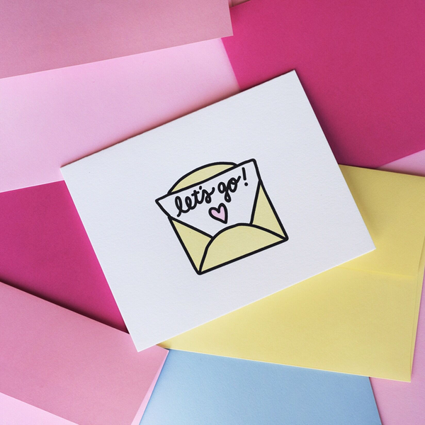 White greeting card with a yellow, black and pink graphic. The graphic is a yellow envelope with a paper sticking out with script text "let's go!" and a pink heart. 