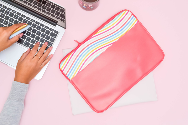 A laptop sleeve sits on a pink background while a woman works on a laptop. 