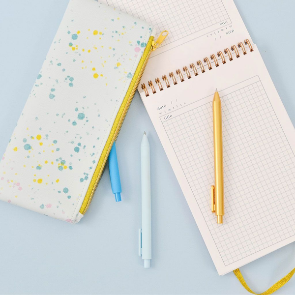 Cute pencil pouch in paint splatter print and a gold zippered top next to a task pad and three jotter pens