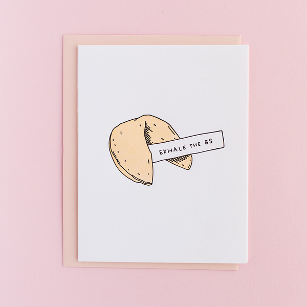 White greeting card with a fortune cookie. The cookie has a piece of paper sticking out of it with the text "Exhale the BS". There is a peach envelope and a light pink background.