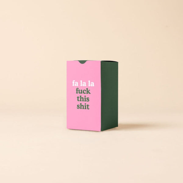 A candle box that is emerald green on three sides and pink on the front side. The pink front side says, "fa la la fuck this shit."