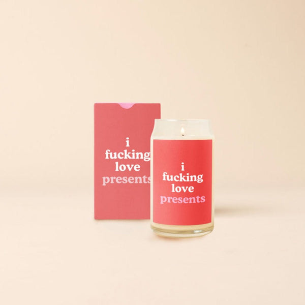 A 12 oz candle with a red decal that says, "i fucking love presents." Candle is displayed with packaging box that is designed the same as the candle decal.