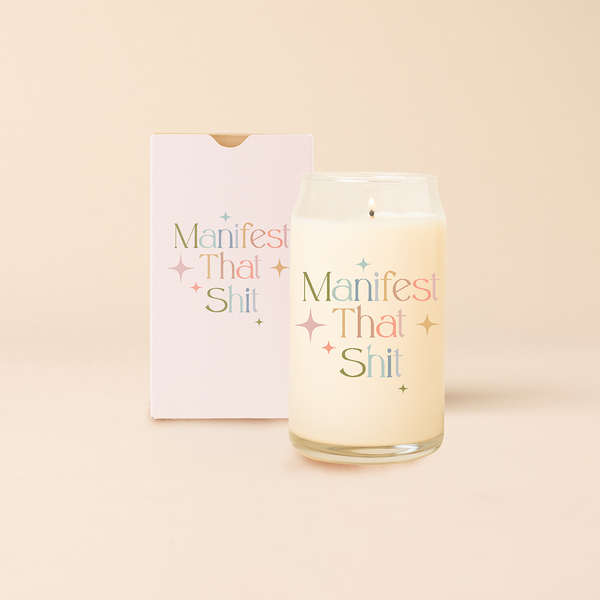 Can glass candle with text that reads "Manifest That Shit" in multi color font, with minimalist sparkle stars surrounding text. Box packaging with the same design sits behind candle.