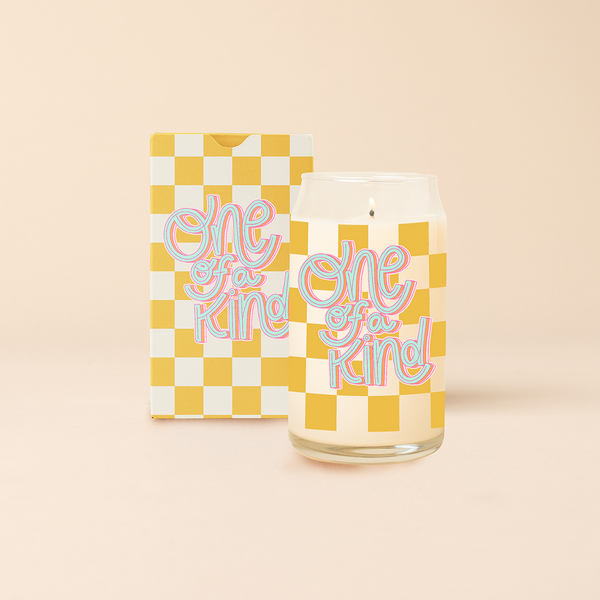 Can glass candle with yellow checkered print wraps around the candle, with text that reads "ONE OF A KIND" in a blue, curly font . Box packaging with same design sits behind candle.