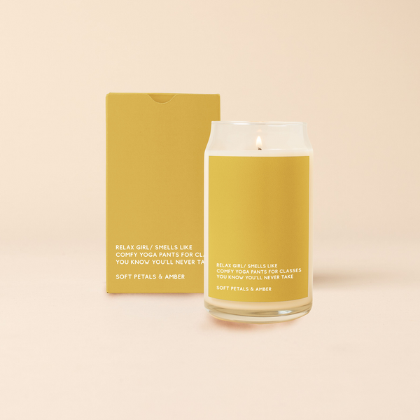 A 16 oz. candle with a mustard yellow colored decal and says, "RELAX GIRL/SMELLS LIKE COMFY YOGA PANTS FOR CLASSES YOU KNOW YOU'LL NEVER TAKE." Scent is Soft Petals & Amber.