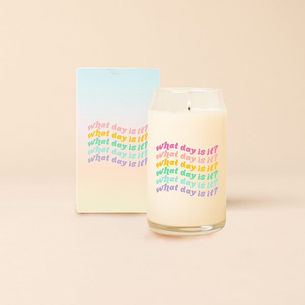Can glass candle with text that reads "what day is it?" in wavy font that repeats 6 times, each in a different color. Box packaging with same design sits behind candle.