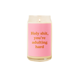 A 16 oz. candle with a pink decal that says "HOLY SHIT YOU'RE ADULTING HARD."